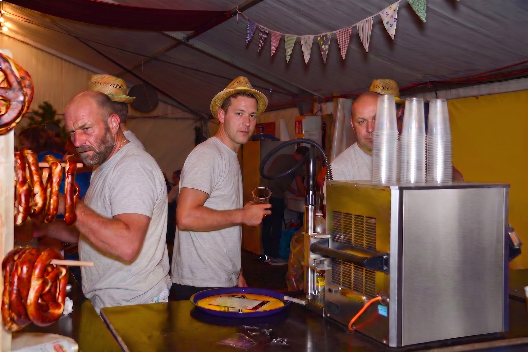 Drei Helferinnen und Helfer in einheitlichen T-Shirts und Strohhüten hinter der Bar einer Festwirtschaft