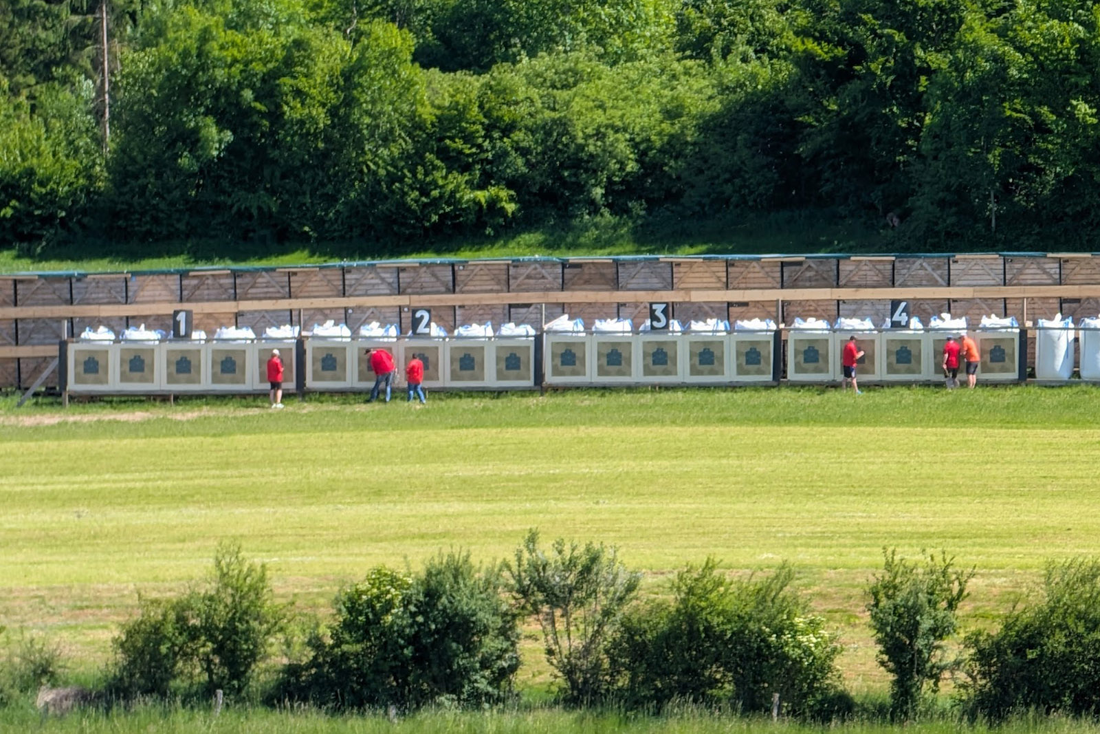Weite Aufnahme des 300m-Scheibenstands mit nummerierten Blocks 1–6, rote Helfer beim Zeigen, Waldkulisse